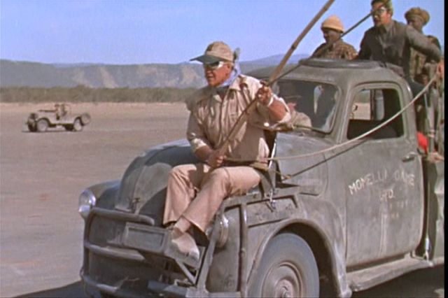 John Wayne from the 1969 movie Hatari. A seat is strapped to the front hood of a pickup truck and John Wayne is sitting in it with a long pole. He looks so cool