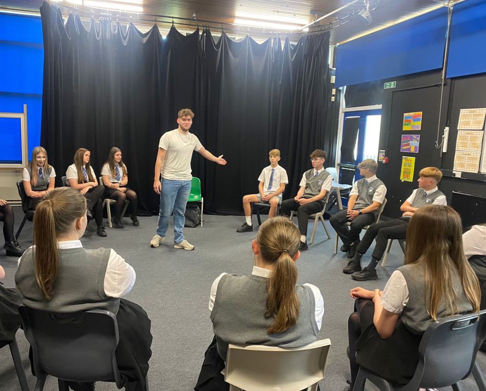 The actor stands in a circle of chairs filled with students. 