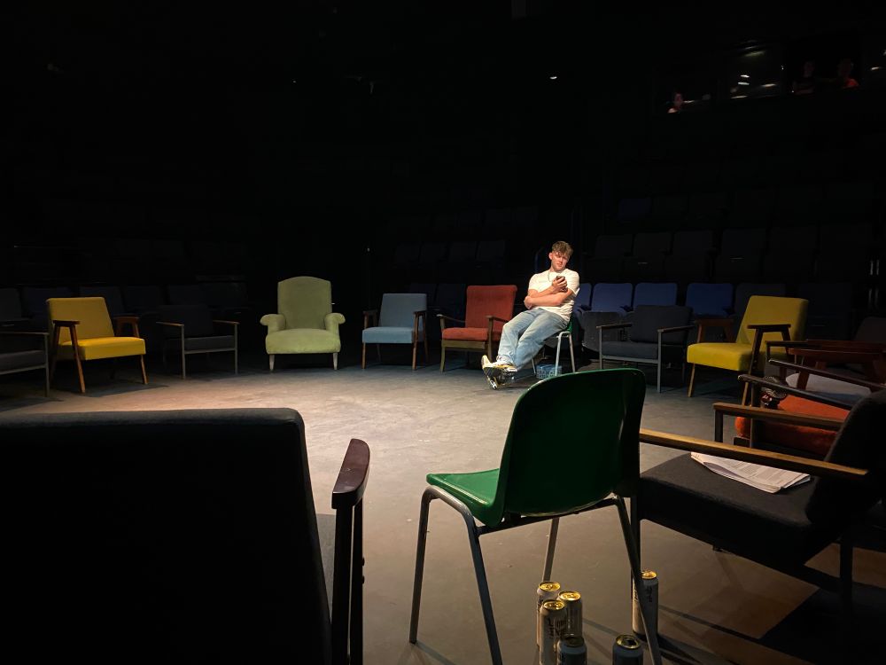 An eclectic mix of chairs arranged in a circle. One has a pile of beer cans beneath and an actor sits scrolling through his phone on another. 