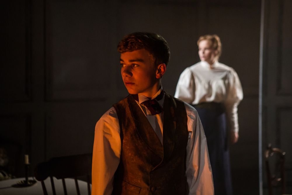 A boy lit by fire light with a Victorian governess in the background from THE TURN OF THE SCREW
