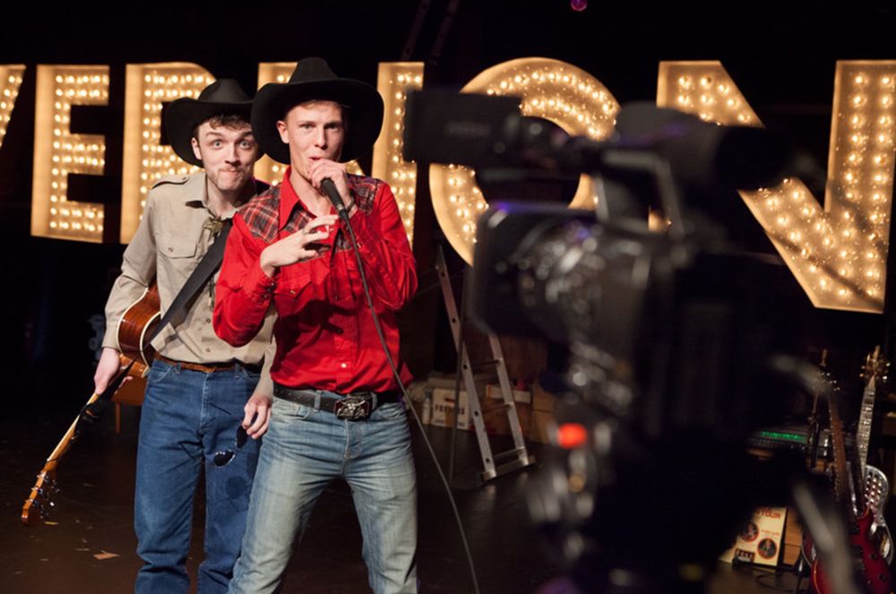 Two men in cowboys hats talking into a video camera stood in front of a giant illuminated sign reading ‘VERNON’ from a production of Vernon God Little. 
