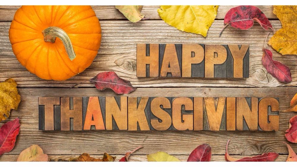 The words Happy Thanksgiving in a cutout style on a wooden background. Fall leaves around and a small pumpkin at top left.