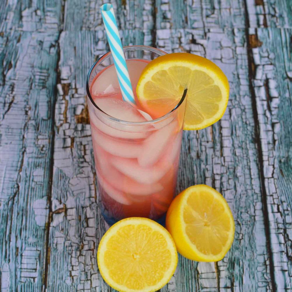 Glass of iced pink lemonade on a grey wooden table, sliced lemon on table next to glass. 