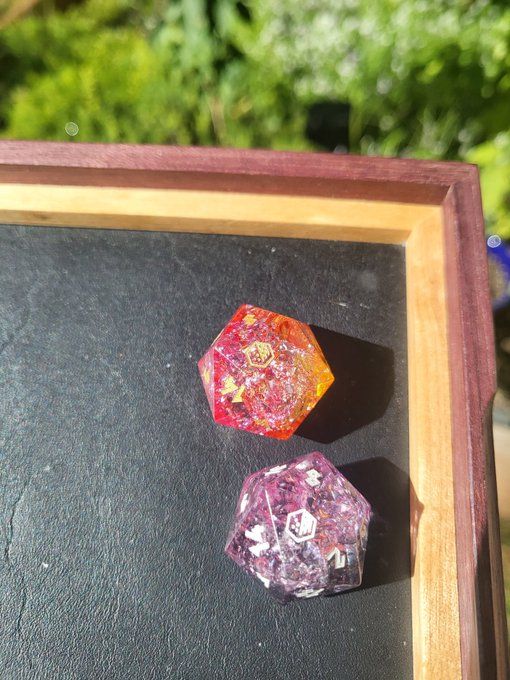 Two twenty sided dice in a dice tray. One is pink and orange, the other lavender and black