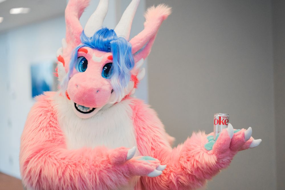 A pink dragon fursuiter holds up a can of diet coke

TAGS: #fursuit #furtographer #anthrocon #fursuitfriday #dragon #AC2025 
