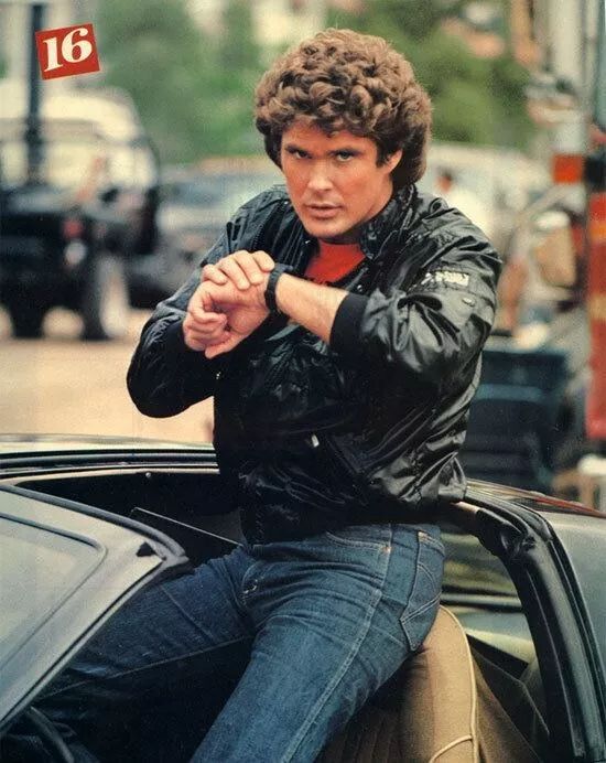 David Hasslehoff in Knight Rider wearing a black Members Only jacket and blue jeans, he's speaking into a watch on his wrist, he's halfway out of the car's roof, which is a semi-convertible.