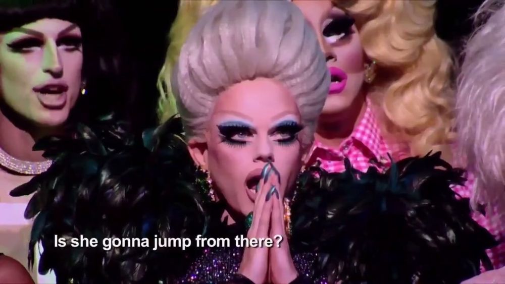 A drag queen with her hands covering her mouth. A subtitle says "Is she gonna jump from there?"