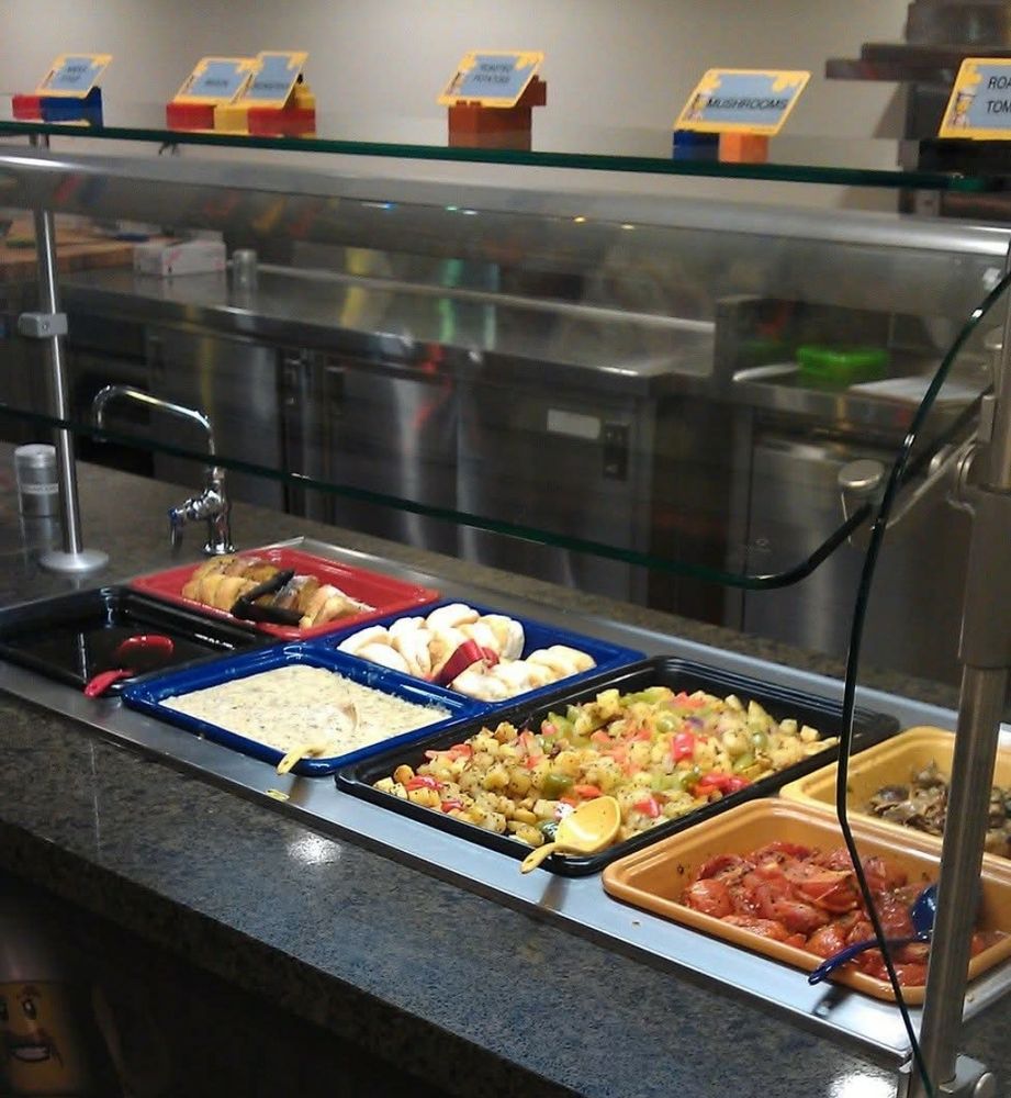 Buffet counter with several hot trays and a sneeze guard at Bricks Family Restaurant in the Legoland Hotel in Carlsbad, California. Offerings include french toast, syrup, American biscuits, white sausage gravy, breakfast potatoes with green and red bell pepper, sauteed mushrooms, and cooked Roma tomatoes. Each item has either a plastic serving spoon or tongs in it.