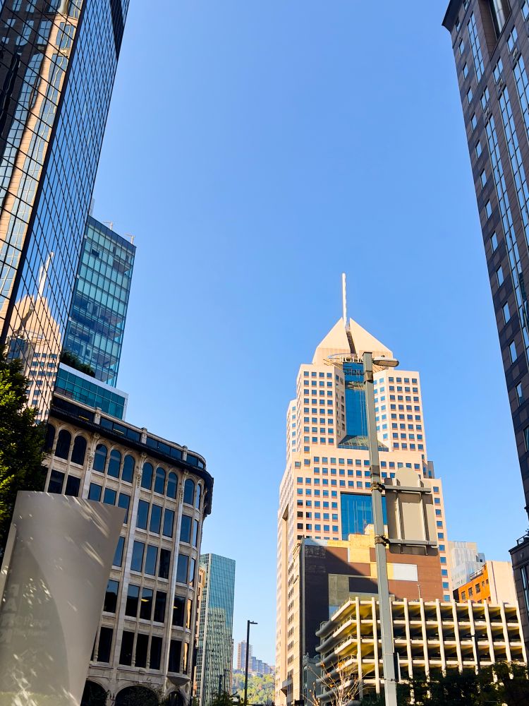 Cityscape featuring tall buildings and a clear blue sky. Notable structures include a modern skyscraper with a geometric design and reflection on glass surfaces, surrounded by other urban architecture.