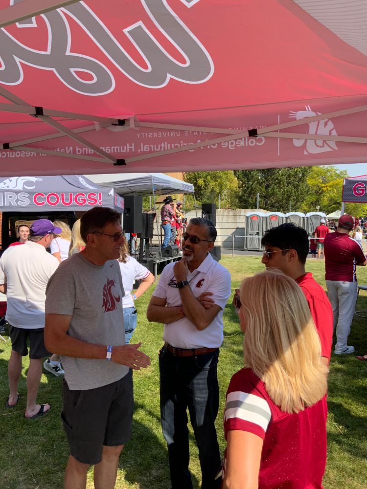 4 people stand talking under a tailgate tent with a band playing behind them.