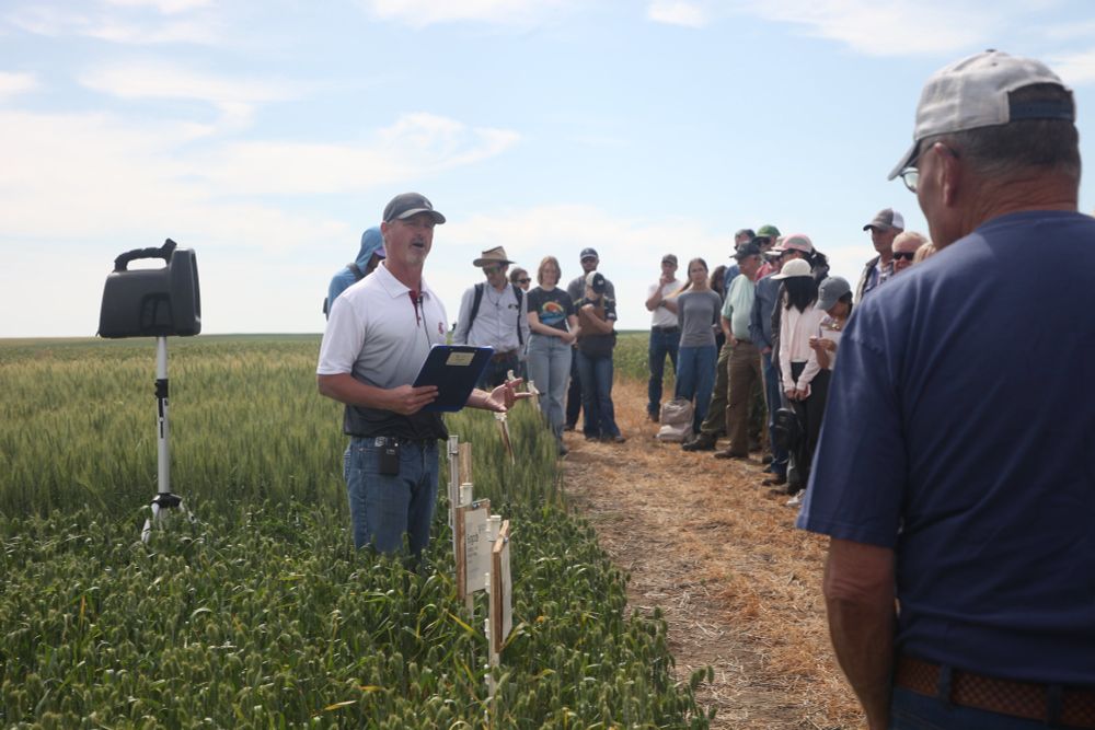 A person stands at the edge of a field, holding a clipboard and gesturing while speaking. Next to the speaker is a large group of people.