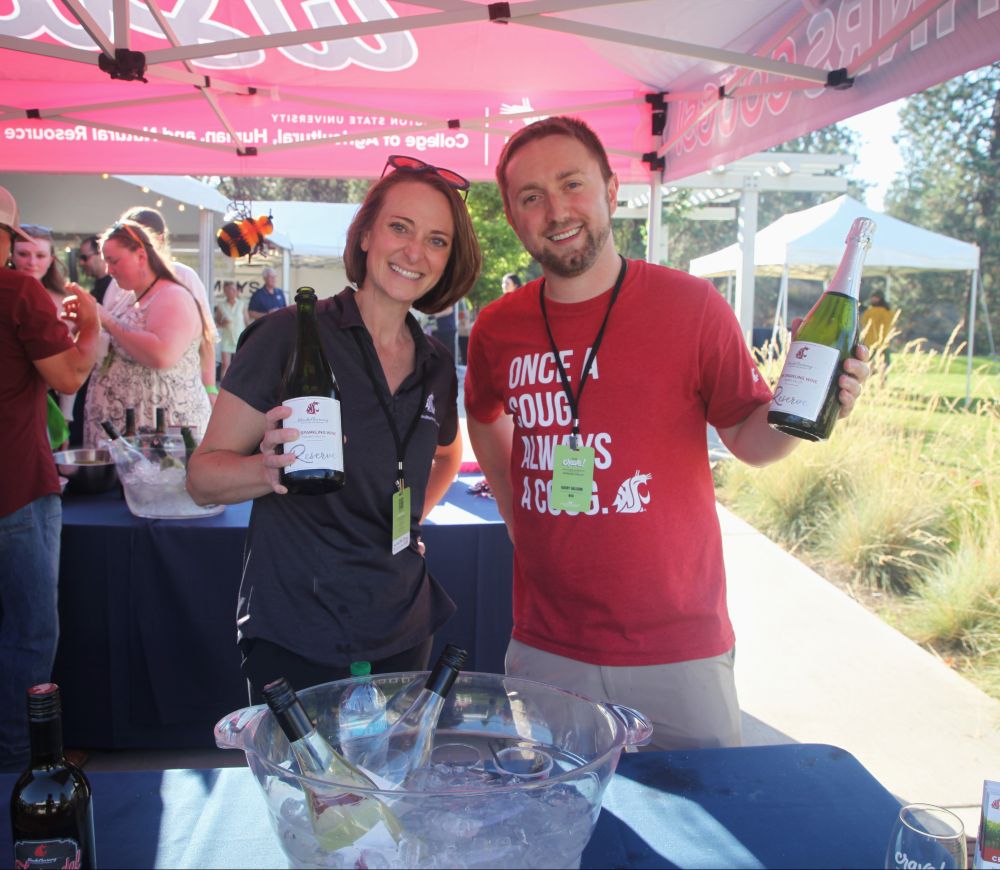 Two people stand behind a table, each holding a bottle of student-made wine. In a bowl of ice in front of them are two more bottles of wine.