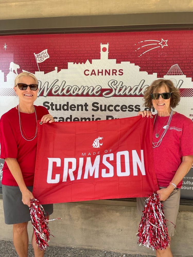 Two people hold pom poms and a banner that reads "Made of Crimson" with the WSU cougar head logo above the words. Behind them is a sign that reads "CAHNRS Welcome Students! Student Success and Academic Programs."