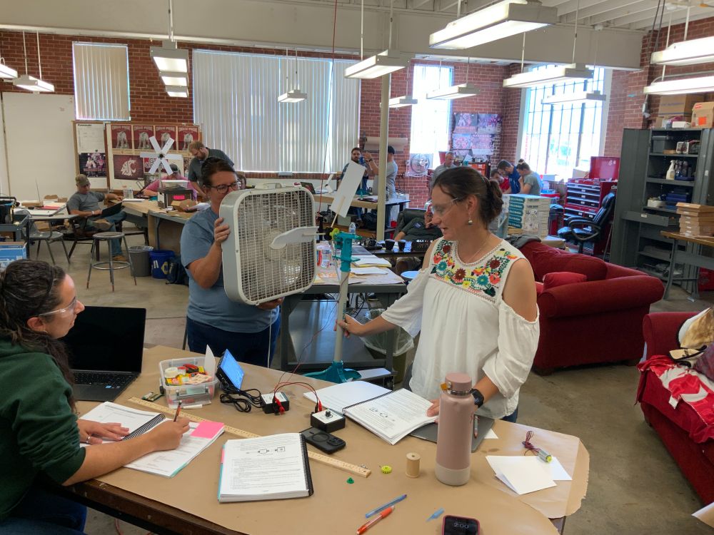In a large room, 1 person holds a box fan up near a windmill, spinning the arms of the windmill. 2 other people talk about the readings they're getting from equipment attached to the windmill. In the background, several other groups are working on the same project.