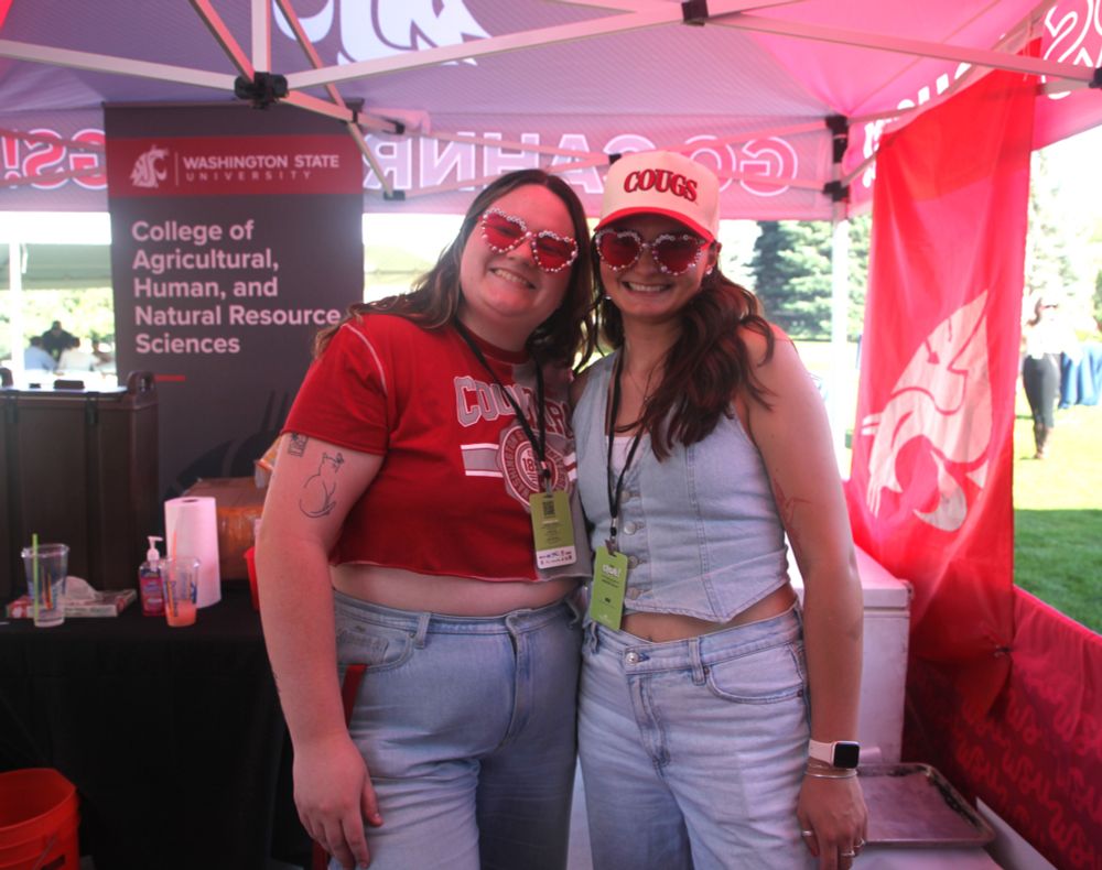 Two people stand together under a WSU-themed tent wearing WSU cougars gear and smiling. Behind them is a banner with the WSU Cougar head logo and the words "Washington State University College of Agricultural, Human, and Natural Resource Sciences." A flag with a WSU cougar head also hangs behind them.