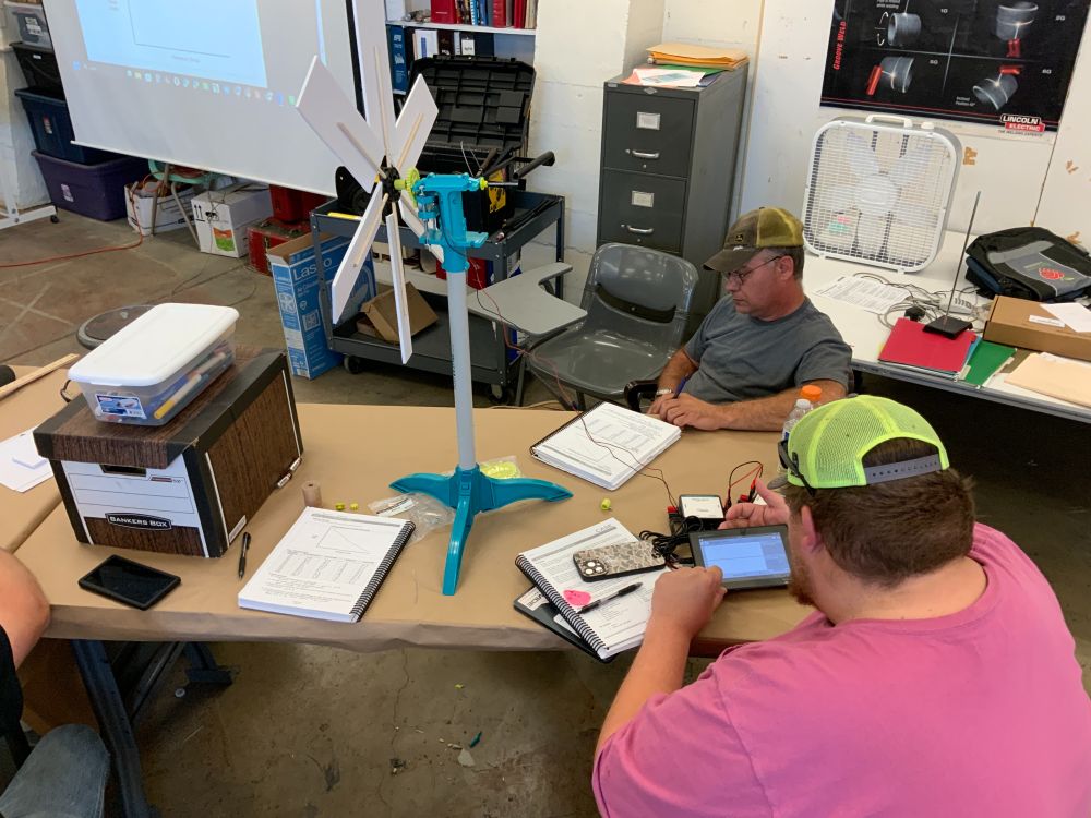 2 people sit at a table, which has a small windmill sitting on it. One person is looking at a screen on a device attached to the windmill.