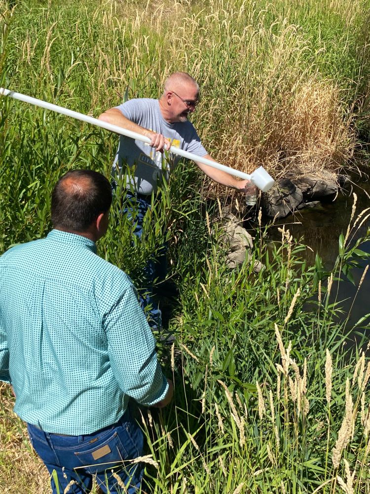 A person stands next to a creek holding a long stick with a white cup attached on one end. He's pouring water from the cup end into another a glass container. A second person watches nearby.