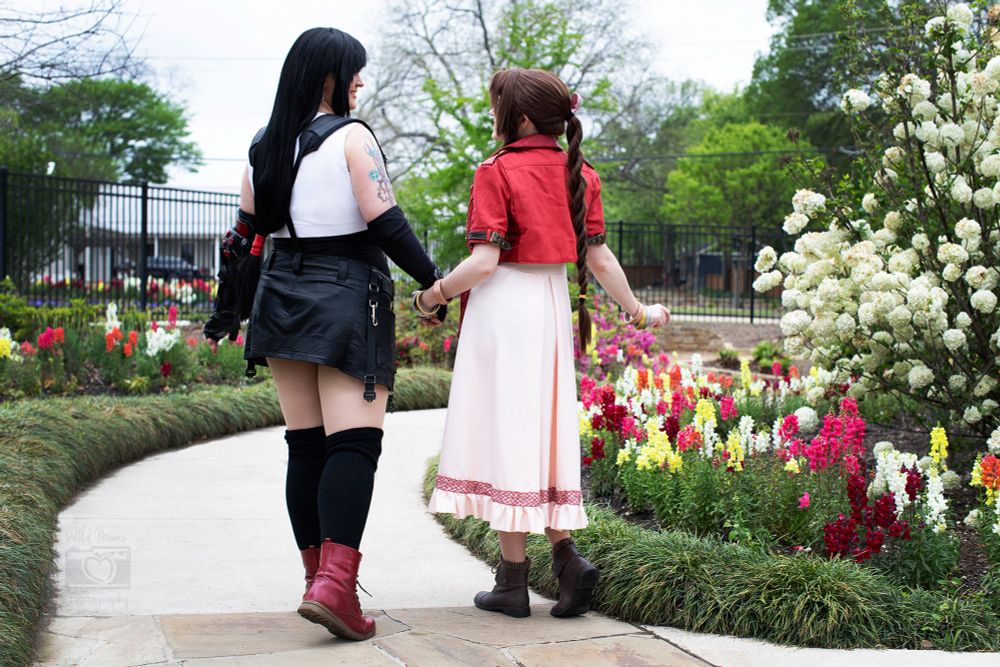A photo of two cosplayers dressed as Tifa and Aerith from FF7 walking through a garden and holding hands.