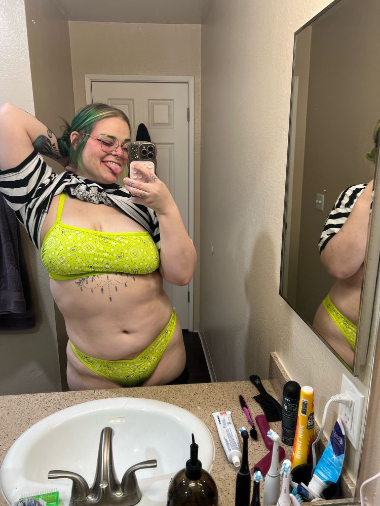green haired model flashes a lime green bra and panty set from the front in the mirror