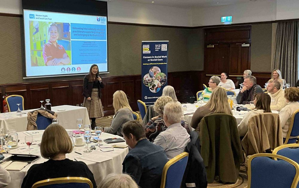 A woman presents information to an attentive audience seated at round tables in a conference room with a projection screen and banner behind her.