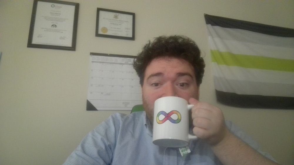 Photo of Drew sipping tea at their desk with a mug bearing the Divergent Financial Planning rainbow infinity logo. Behind Drew are their degrees, wall calendar, and a portion of an agender flag.