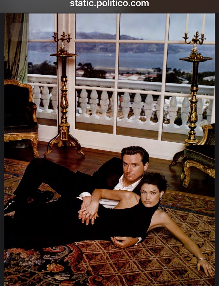 Gavin Newsom is lying on the floor with Kimberly Guilfoyle on an orate orangish rug with tall golden candlesticks and ornate white railing in the background. The photo is from when they were married and before she put on her MAGA face. 