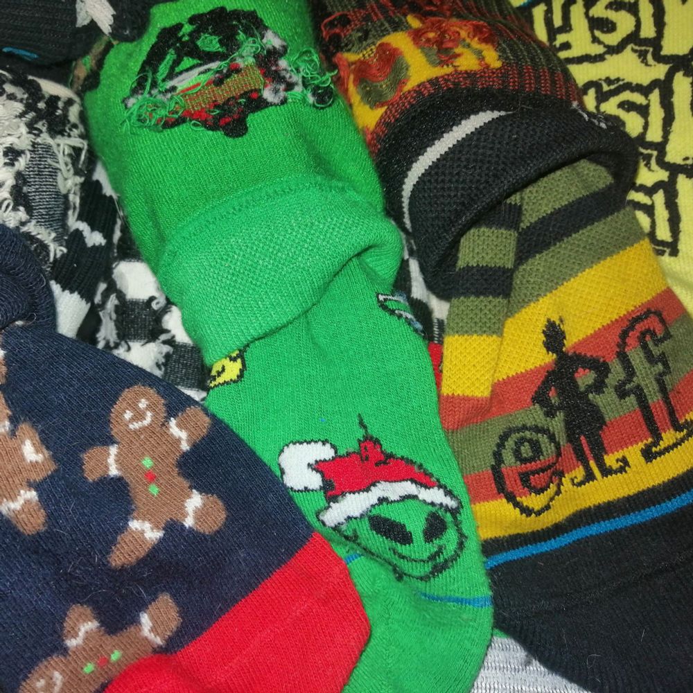 En bunke farverige sokker med forskellige mønstre: en grøn sok med et julemotiv af et lille alien-ansigt med nissehue, en sok med stribede reggae-farver og en silhouet-figur, en sok med små peberkagemænd, samt andre mønstrede sokker i sort, hvid og gule nuancer