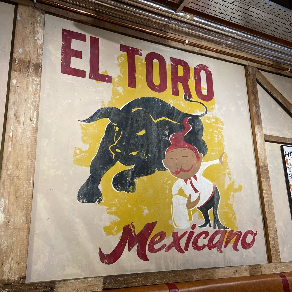 A fake poster on the wall in a burrito restaurant in Berkshire, England. It shows a bullfighter and a large black bull, and the title 'El Toro Mexicano'.