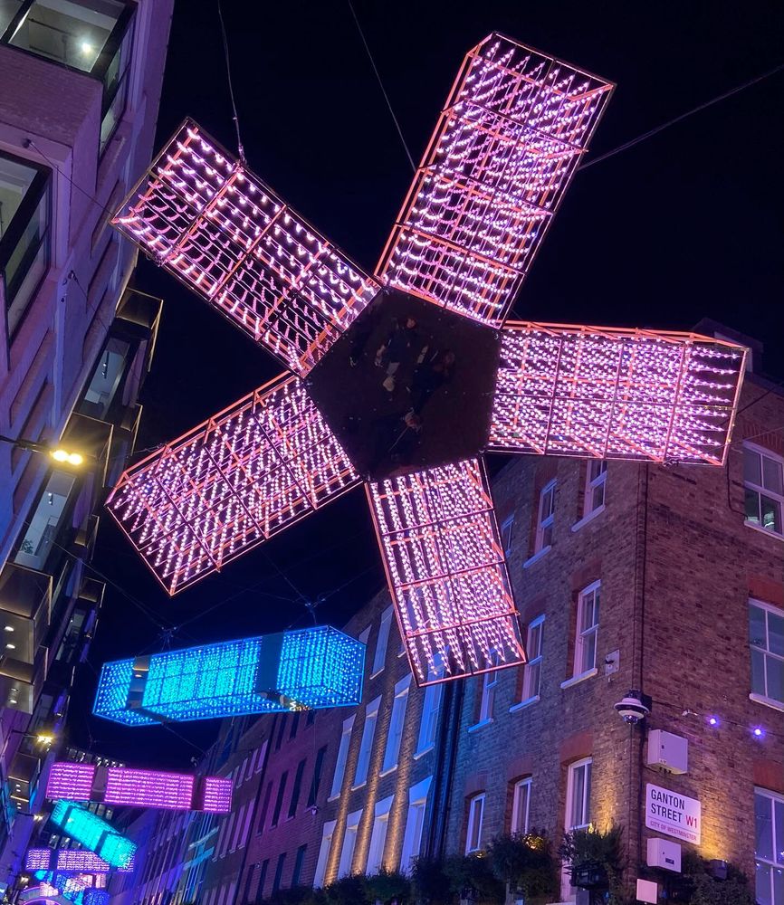 Christmas lights suspended above Carnaby Street in London - huge stylised stars and Christmas crackers, made of square tubes of lights that look a bit like stickle bricks. In this picture, a pink star, and then blue, purple, and green crackers.