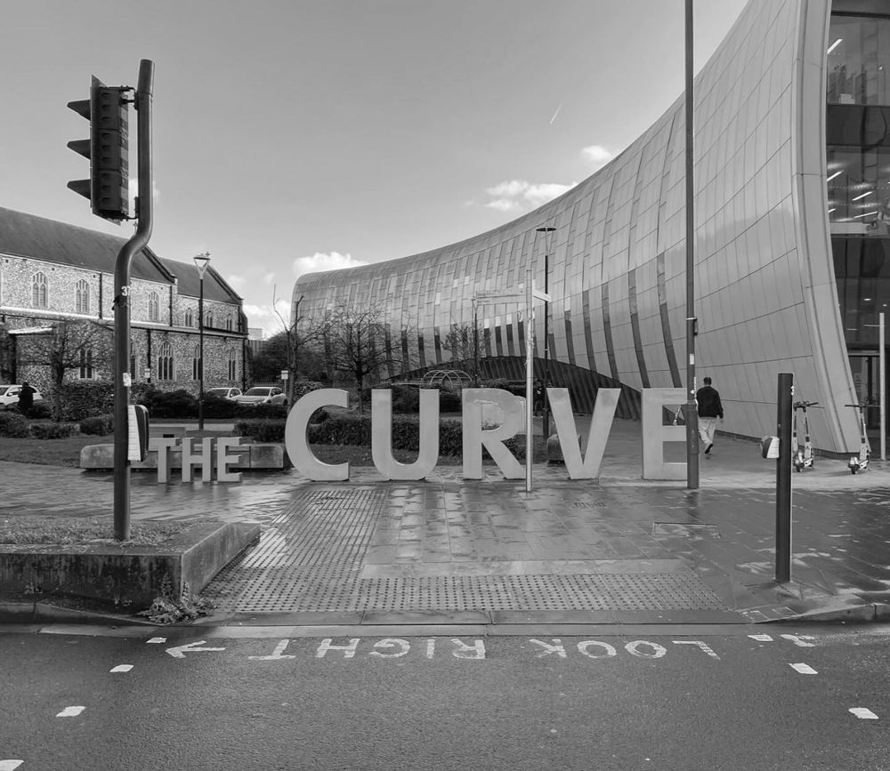 Black-and-white photograph - The Curve in Slough. A curved three-story building with curved metal cladding, with a large sign next to it saying 'The Curve' in freestanding letters that are about four feet high.