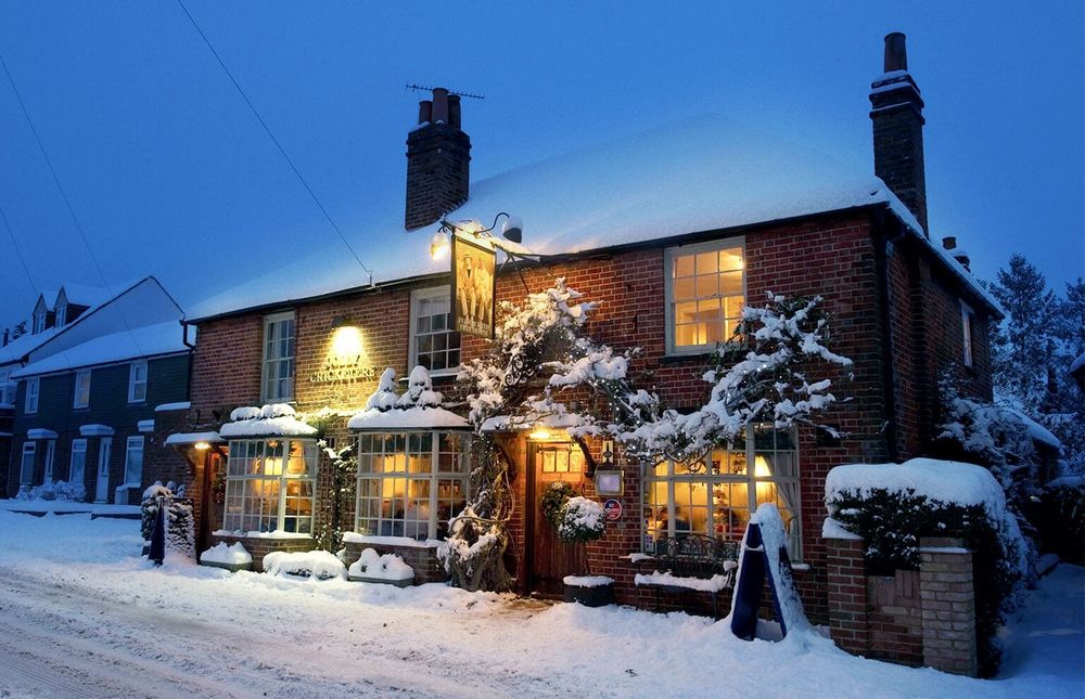 An old countryside pub covered in snow, at dusk with lights on inside. It looks cosy.