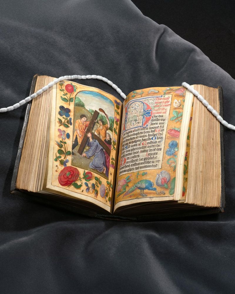 Een middeleeuws handschrift, opengeslagen op een kussen, met op één pagina een rijkversierde tekst en op de andere pagina een afbeelding van Jezus die het kruis tilt terwijl omstanders hem lastigvallen.