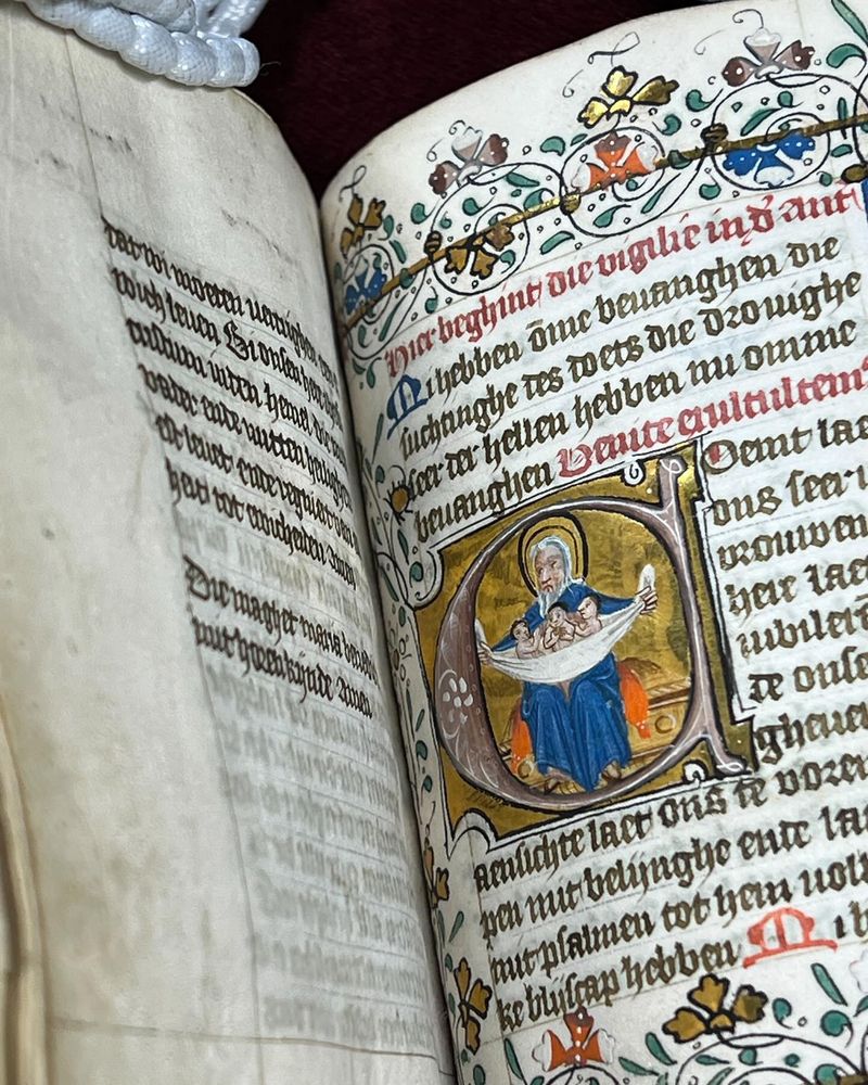 Een pagina uit een middeleeuws manuscript met een rijk versierd initiaal waarin een man een doek met drie baby's vasthoudt. De rand van de pagina is versierd met gekleurde en gouden bloemen. 