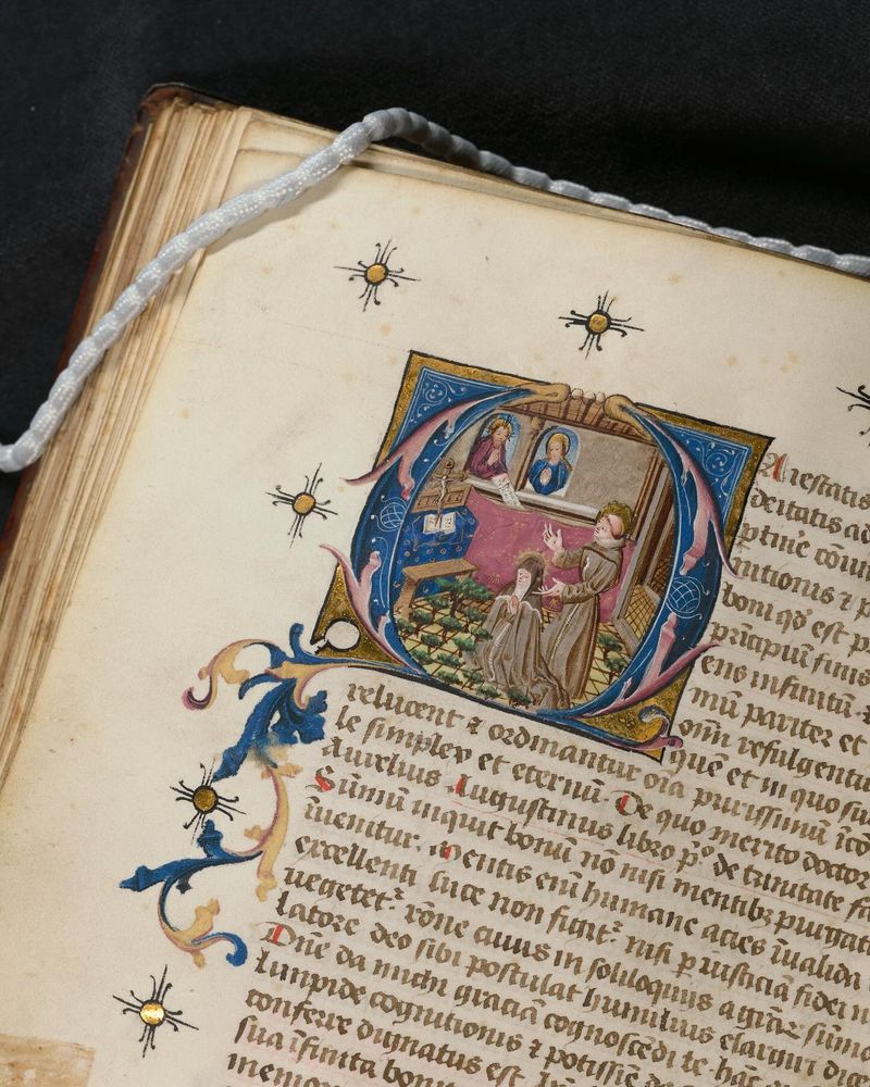 Een rijk versierd initiaal in een middeleeuws handschrift. Getoond word een knielende non, Sint Coleta, achter haar een man met een aureool, Sint Franciscus. Op de muur hangen afbeeldingen van Jezus en Maria. De afbeelding heeft een rand met blauw en goud die uitloopt in een soort bladerkrans langs de tekst. 