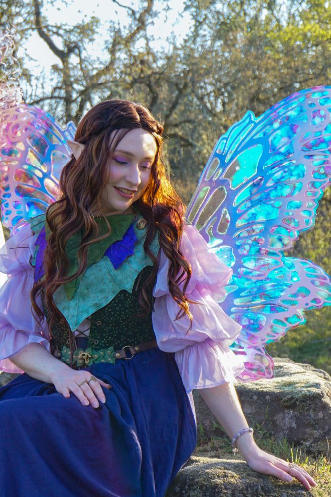 Fairy sitting upon a rock with spread out wings shining blues, purples, and pinks. She looks down smiling softly with a green and purple leaf cowl around her neck. 