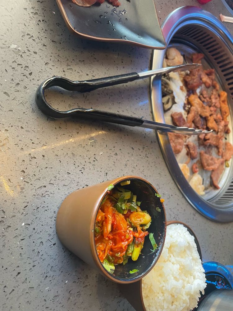 Picture from probably a Korean BBQ restaurant.

There's a recessed circular stove top covered in strips of brown meat. a pair of tongs are resting on the table. There is a bowl of white rice and a different bowl of brightly colored orange and green veggies. There might be a red sauce on the veggies