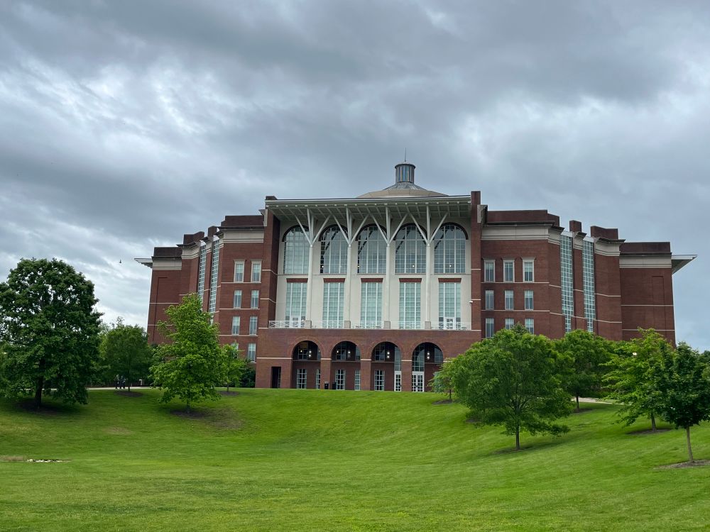 Picture of a University of Kentucky library building with grass and trees in front and dark clouds above