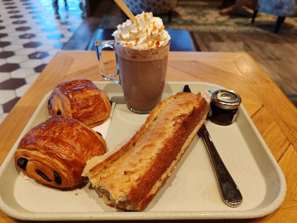 Plateau avec deux pains au chocolat, une demi baguette de pain, un couteau, de la confiture de framboise, du beurre demi-sel et un verre de chocolat Viennois.
