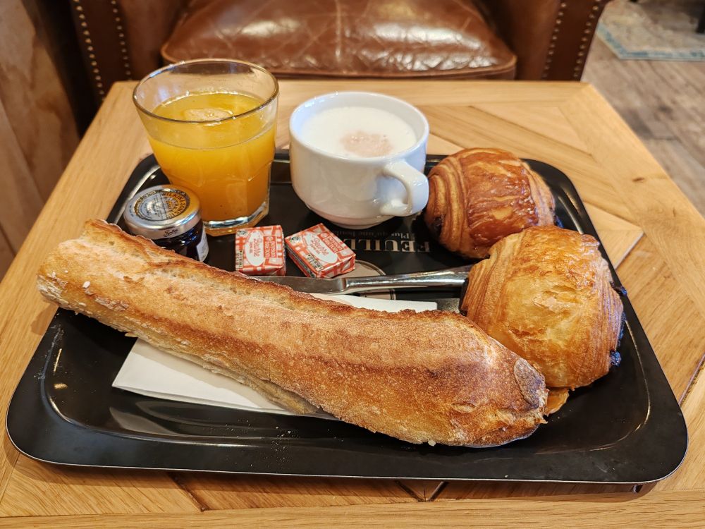Plateau avec deux pains au chocolat, un petit pot de confiture framboise, une demi baguette, deux plaquettes de beurre demi-sel, une tasse de chocolat chaud et un verre de jus d'orange.