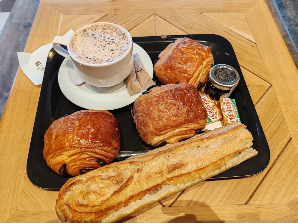 Plateau avec trois pains au chocolat, une demi baguette, deux plaquettes de beurre demi-sel, un petit pot de confiture de myrtilles ainsi qu'une tasse de chocolat chaud.