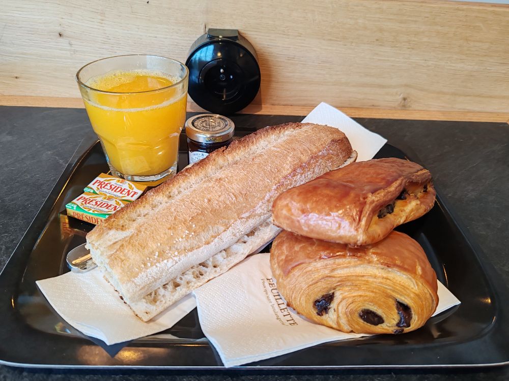 Plateau avec : un verre de jus d'orange, une demi baguette de pain, un petit pot de confiture de framboise, deux petites plaquettes de beurre demi-sel, un pain suisse et un pain au chocolat (ou chocolatine pour les gens du sud)