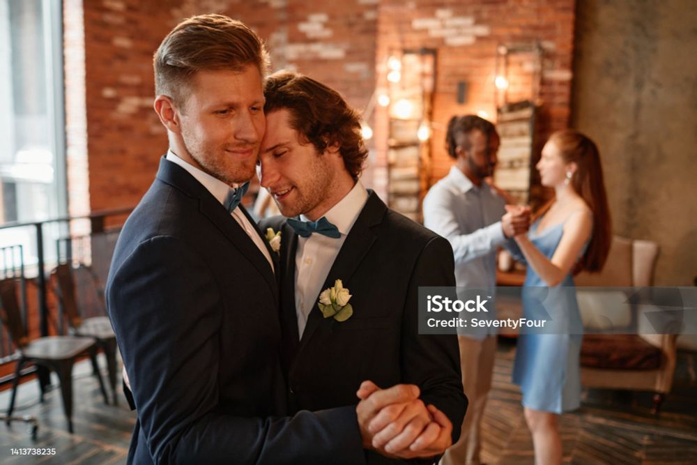 Two men at a wedding dancing. They are clearly the two getting married. They are wearing grey tuxes and blue bowties with light yellow flowers in their lapells. One man is roughly six inches or 20 centimeters taller than the other. The tall man is blond with swoopy hair on top and tight on the sides, the shorter man is brunet with curly hair. The short man is leaning his forehead against the tall man's cheek