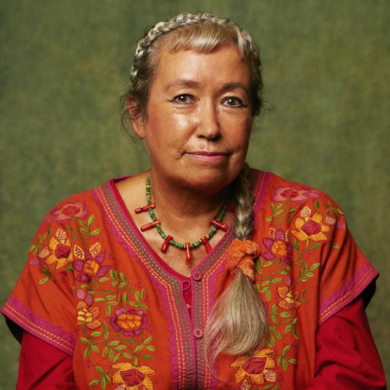 The photograph of Pascale Petit taken by Derrick Kakembo shows Pascale wearing a red top embroidered with yellow and pink flowers with green leaves. The background is green. 
