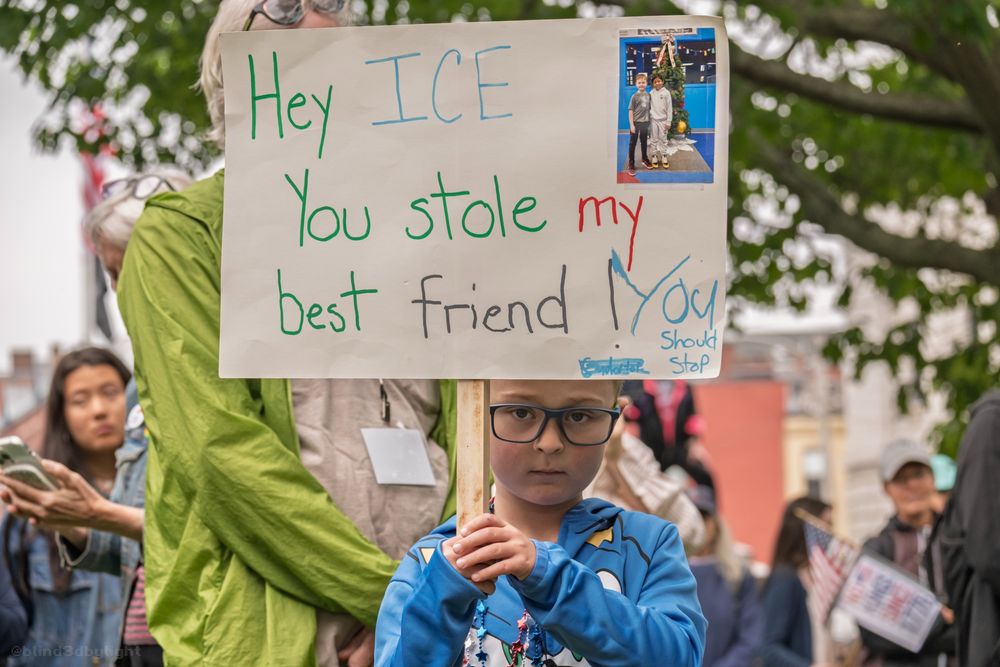 Photo of a bespectacled young boy at the NO KINGS protest in Portland, ME. He is holding a sign that reads, "HEY ICE: YOU STOLE MY BEST FRIEND! YOU SHOULD STOP". At the upper right corner of the sign is a photo of the sign-holding boy with his Latino friend, presumably, the best friend who was taken by ICE.