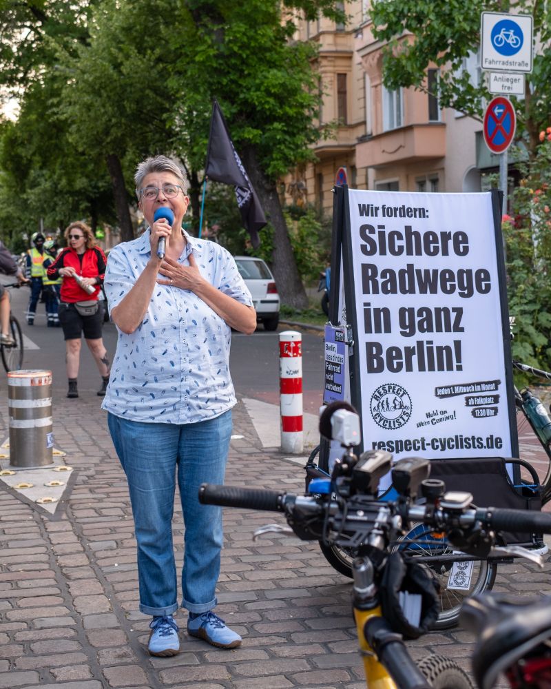 Monika Hermann spricht bei einer Zwischenkundgebung in der Körtestraße in Berlin, neben einem Plakat mit der Forderung nach sicheren Radwegen in ganz Berlin
