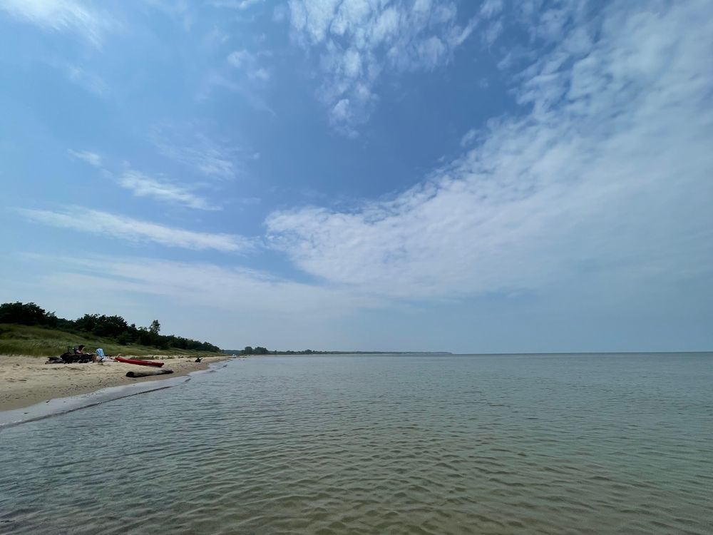 A picture of Lake Huron with a partially cloudy sky looking down the beach as a few people are on shore with a kayak that I took while standing in the water