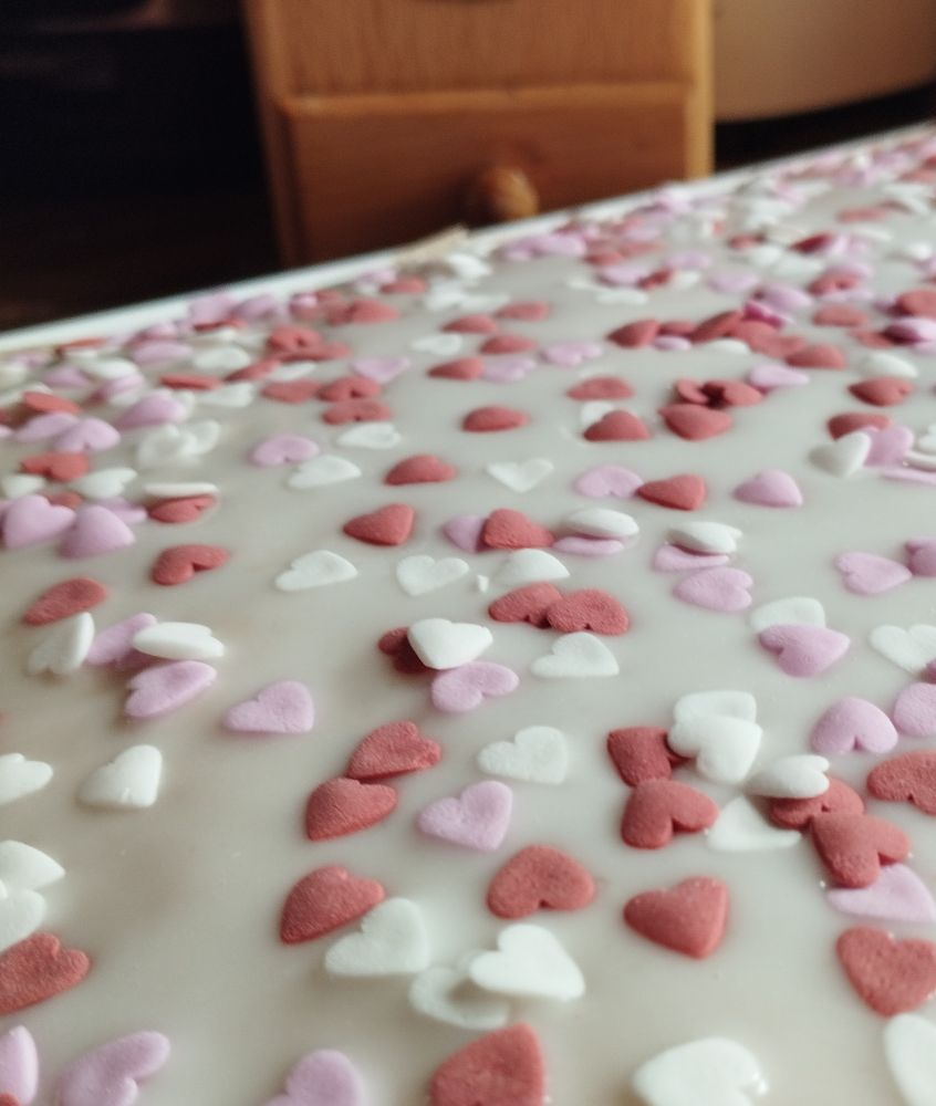 Foto eines leckeren Blechkuchens mit Zuckerguss und rosa und weißen Zuckerherzchen. Im Hintergrund ist ein Teil einer Kaffeemühle aus Holz zu sehen.
