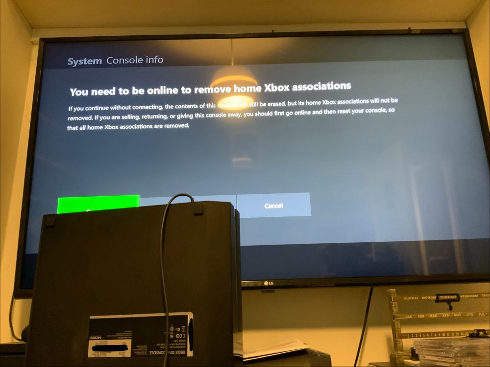 Photograph of a TV partially obscured by an upside-down Xbox One gaming console, with text on screen reading:
"You need to be online to remove home Xbox associations
If you continue without connecting, the contents of this console will still be erased, but its home Xbox associations will not be removed. If you are selling, returning, or giving this console away, you should first go online and then reset your console, so that all home Xbox associations are removed."