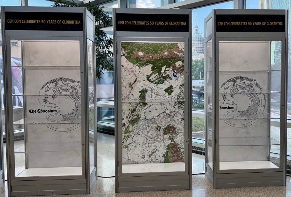 Three glass cabinets emptied of their contents, but with paper still visible above and behind shelves. At top reads “GenCon Celebrates 50 Years of Glorantha.” Two cabinets show the original logo of “The Chaosium”, and central cabinet shows original map of Glorantha.