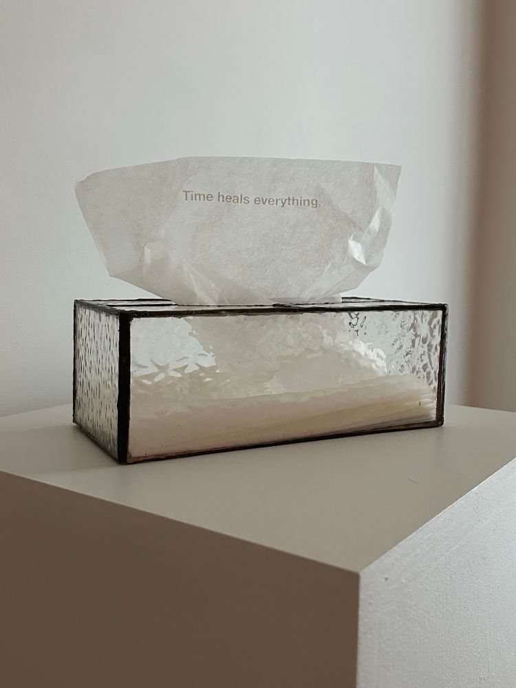 A glass tissue box sits on top of a white plinth. The glass is clear, but has a slight bubbly texture to it. The solder has been patina’d to look aged. The tissue reads, “Time heals everything.” The background is also white and the light comes from the right hand side of the image. 